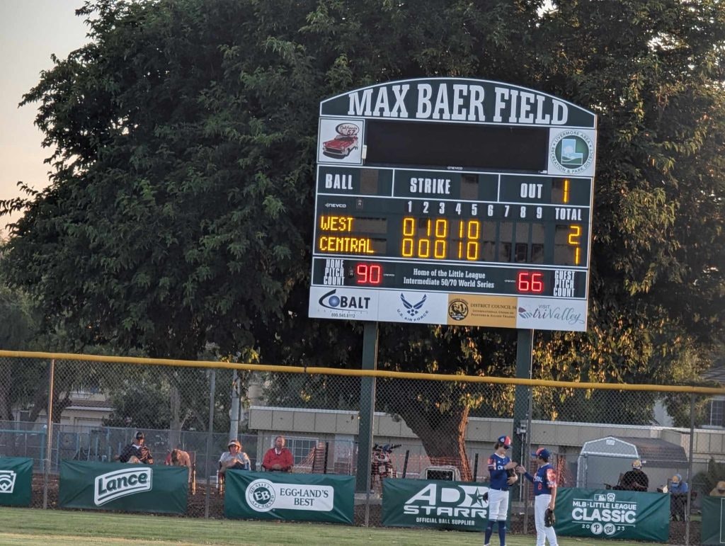 A baseball scoreboard, spectators, and players on the field during a game.