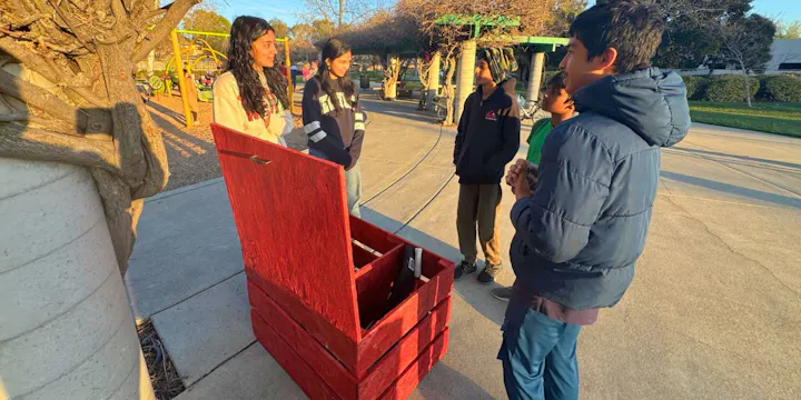A group of children gathers around a red treasure chest in a park, engaging in conversation and enjoying the outdoors.
