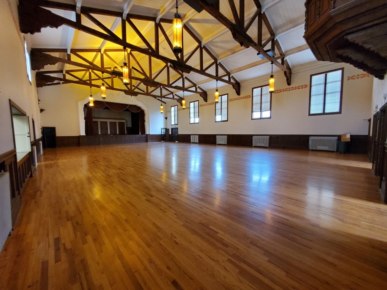 Spacious hall with wooden floors, high ceiling beams, hanging lights, and a stage at one end.