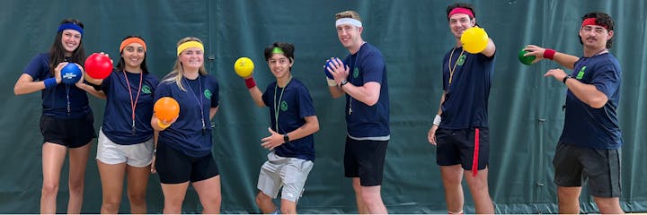 A group of seven people in sports attire holding colorful dodgeballs, wearing headbands and wristbands, pose cheerfully indoors.