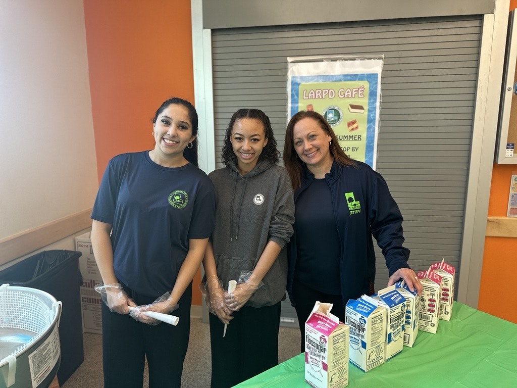 Three staff members stand behind a table with milk cartons and a green tablecloth, promoting the LARPD Café.