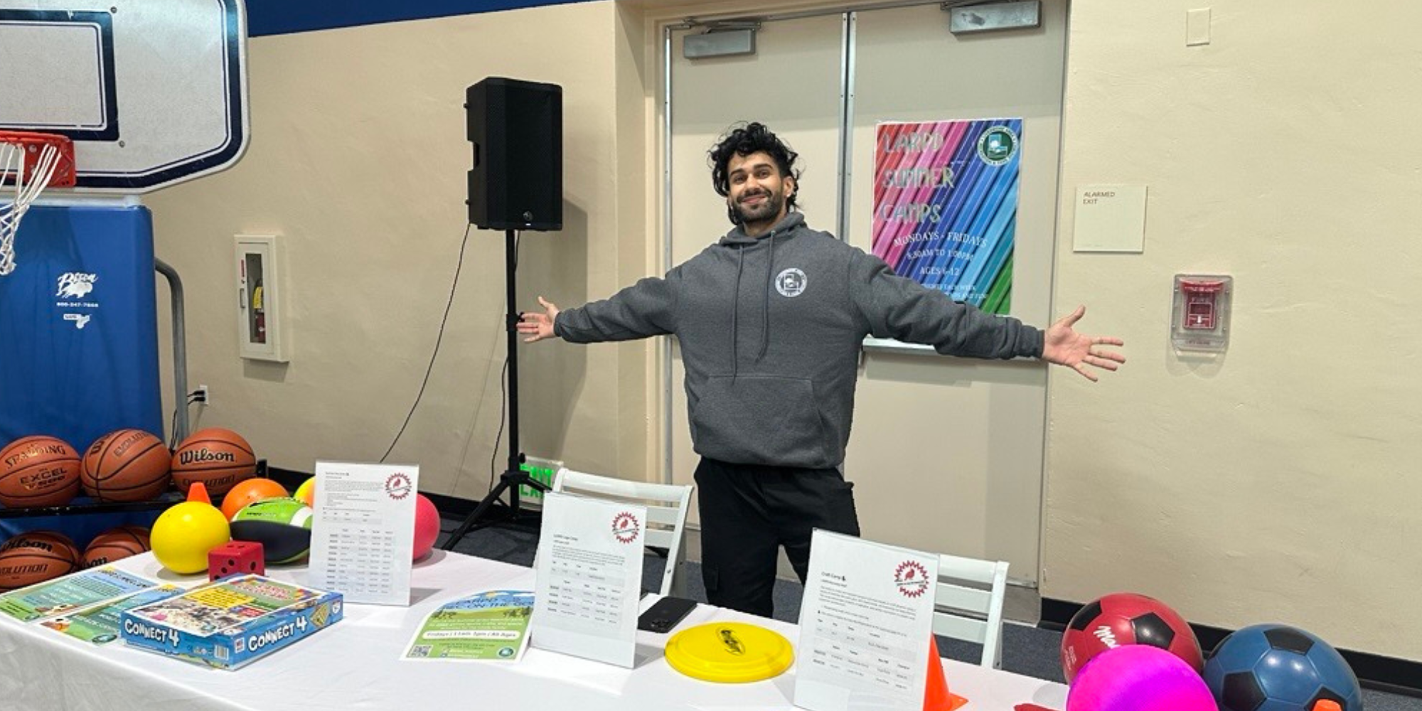 A person stands at a table with games and sports equipment, celebrating or promoting an activity in a gym-like setting.