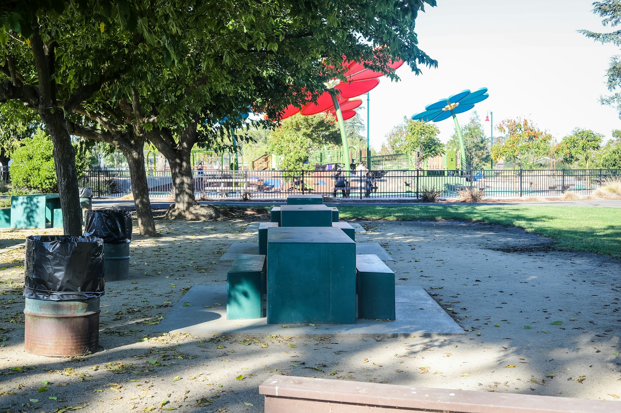 A playground with colorful shades, green play structures, trash bins, and trees on a sunny day.