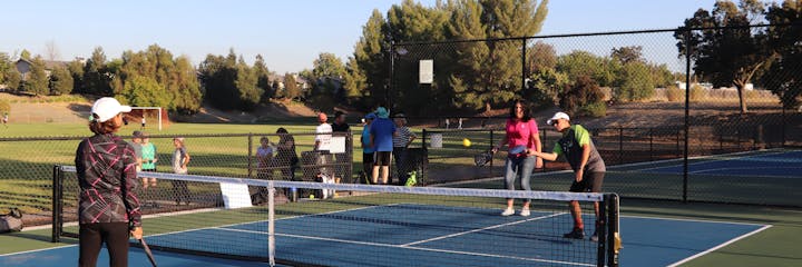 People playing pickleball on outdoor courts, with some spectators watching. Sunny day.