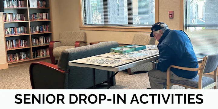 A senior man is working on a puzzle at a table in a library, surrounded by bookshelves, with a sign for drop-in activities.