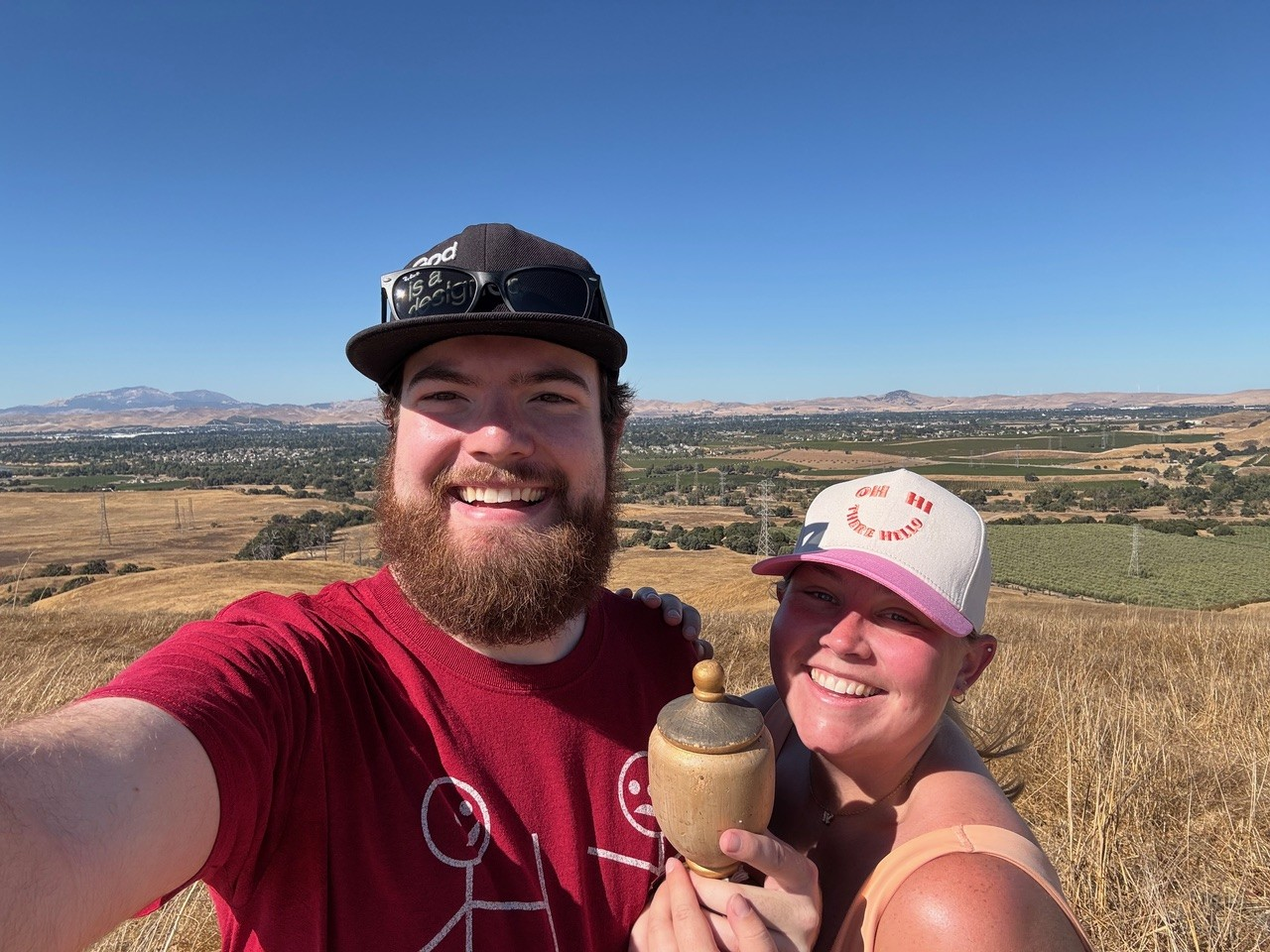 Two smiling people taking a selfie in a field with mountains in the background.