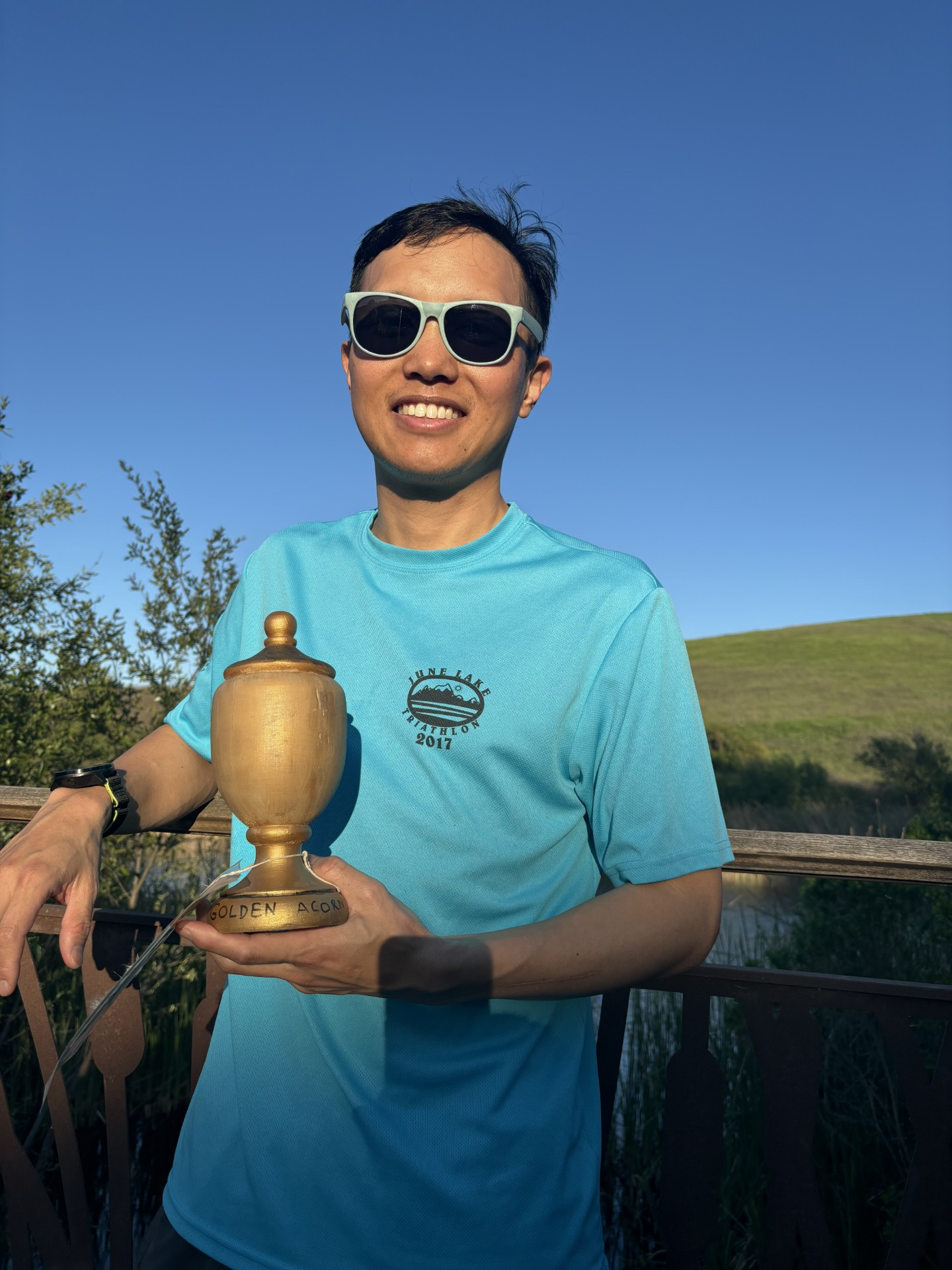Person holding a trophy with "GOLDEN ACORN" text outdoors, wearing sunglasses and a blue t-shirt.