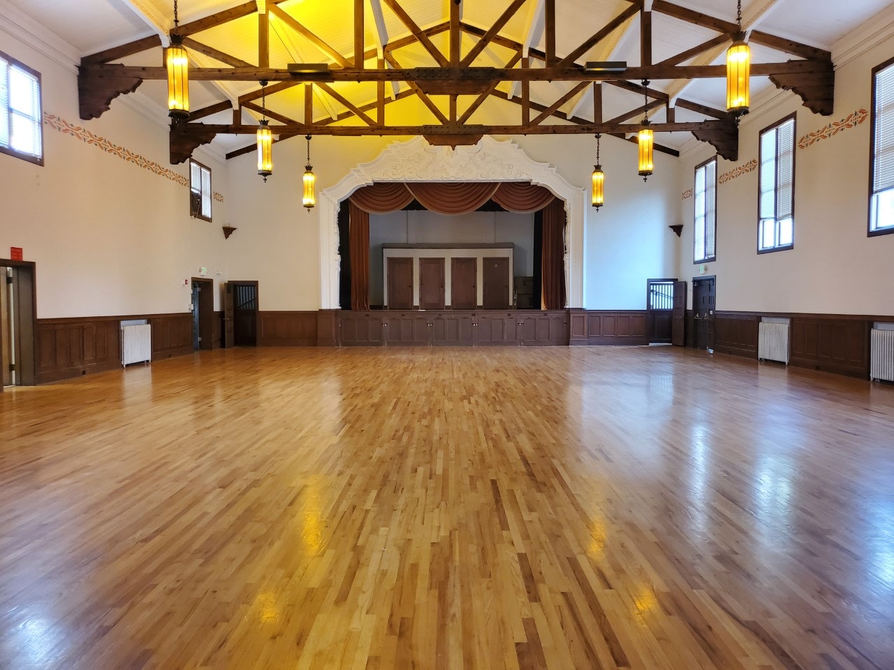 Spacious hall with wooden floors, a stage, high ceiling with beams, and hanging lights.