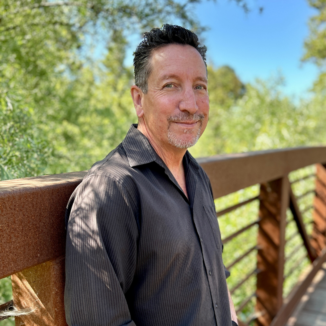 A person in a dark shirt is standing on a wooden bridge with lush greenery in the background.