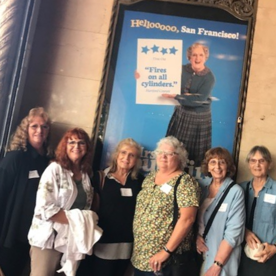 Group of women posing in front of a poster with a cheerful greeting "Helloooo, San Francisco!" and a theatrical review quote.