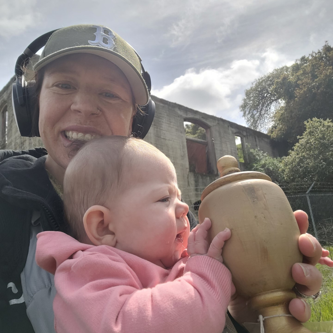 A smiling adult with headphones and a baby holding a wooden object, with a concrete structure and greenery in the background.