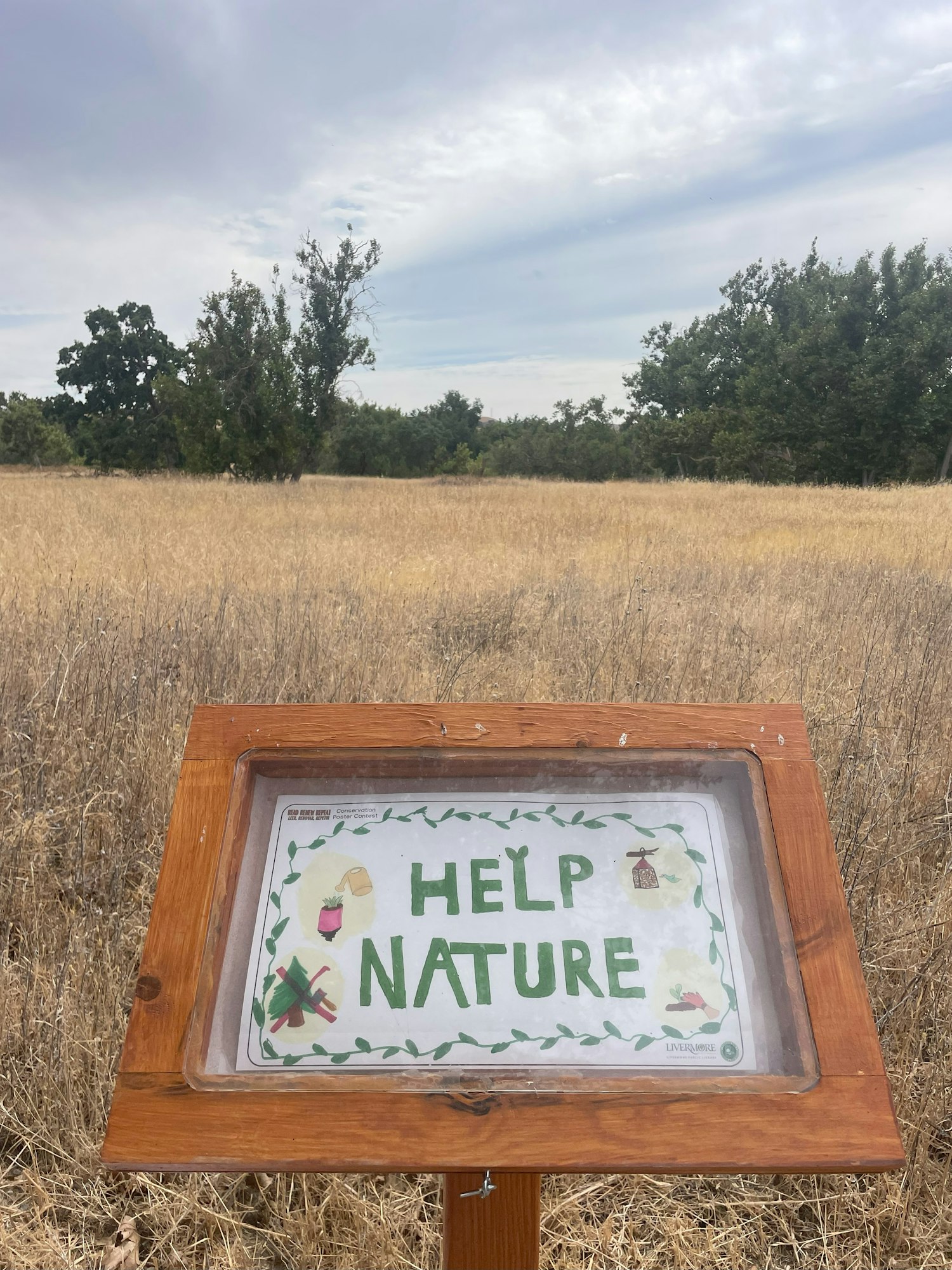 A sign reading "HELP NATURE" in a wooden frame with a dry grass field and trees in the background.
