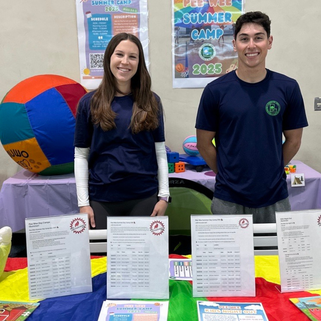 Two camp staff members stand beside tables displaying information about a summer camp, colorful games, and schedules.