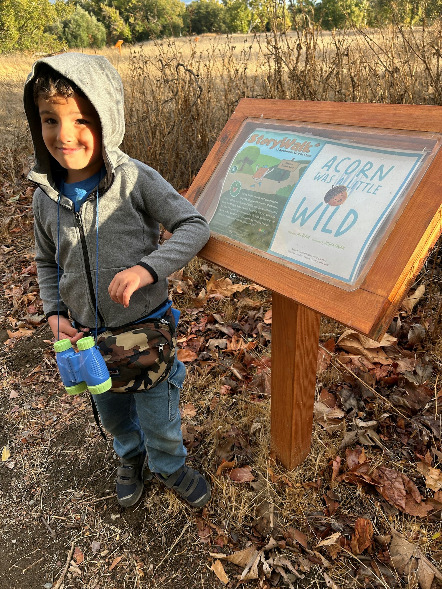Smiling child with a water bottle next to an informational sign on a trail.