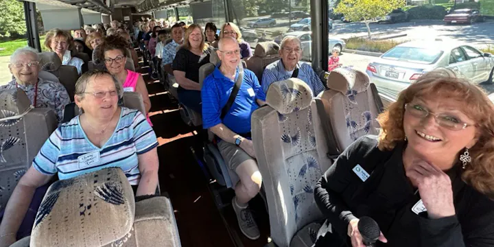 A group of smiling people sitting inside a bus, with a woman at the front holding a microphone.
