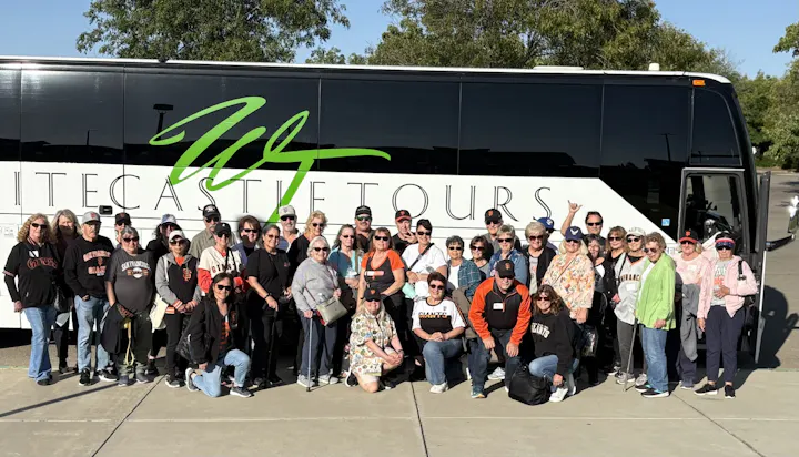 A large group of people, many in San Francisco Giants gear, poses in front of a tour bus, smiling for a photo.