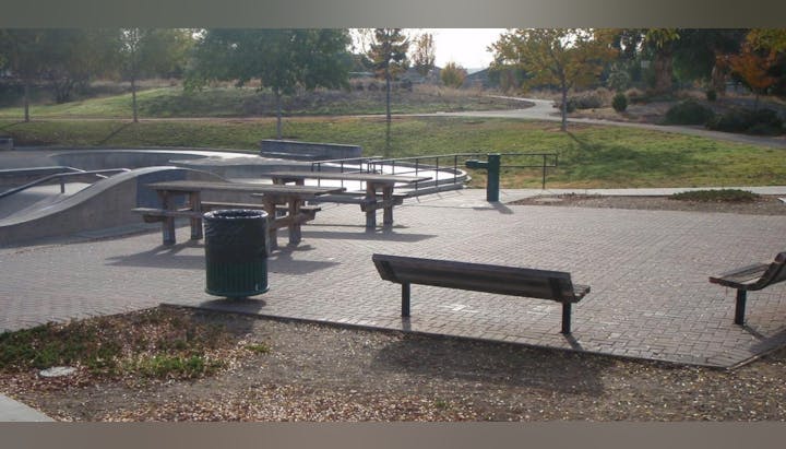 A park with picnic tables, benches, a trash can, and a skate park in the background.