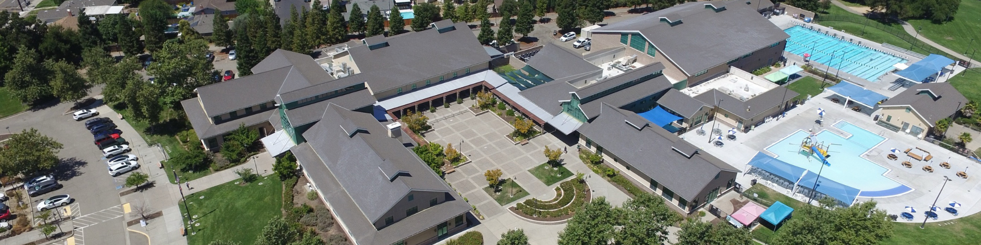 Aerial view of a complex with buildings, parking, pool, and greenery.
