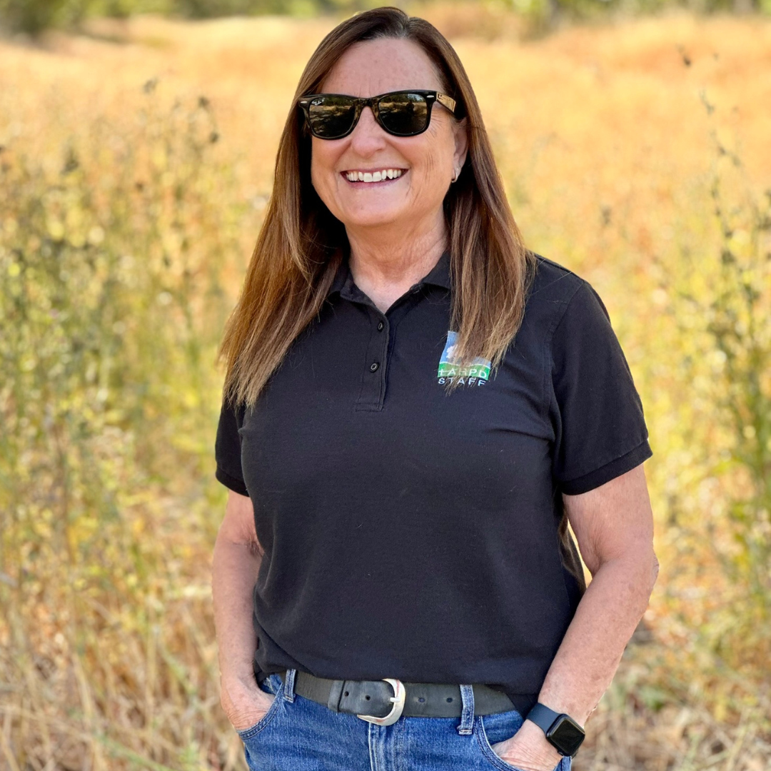 A person in sunglasses smiles outdoors, wearing a black polo and jeans, hands in pockets against a grassy background.