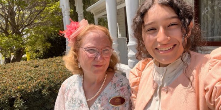 Two women, one older with glasses and flower accessory, and one younger smiling, sitting in a garden setting with a porch.