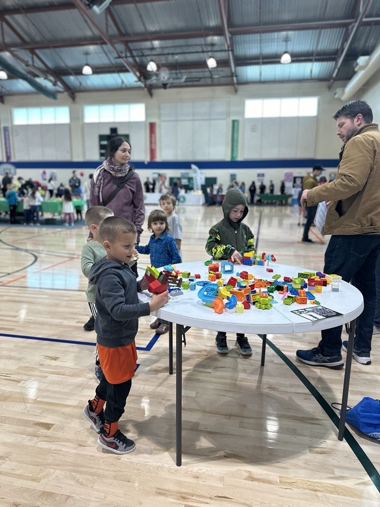 Children play with colorful building blocks on a table in a gym, with adults supervising in the background.