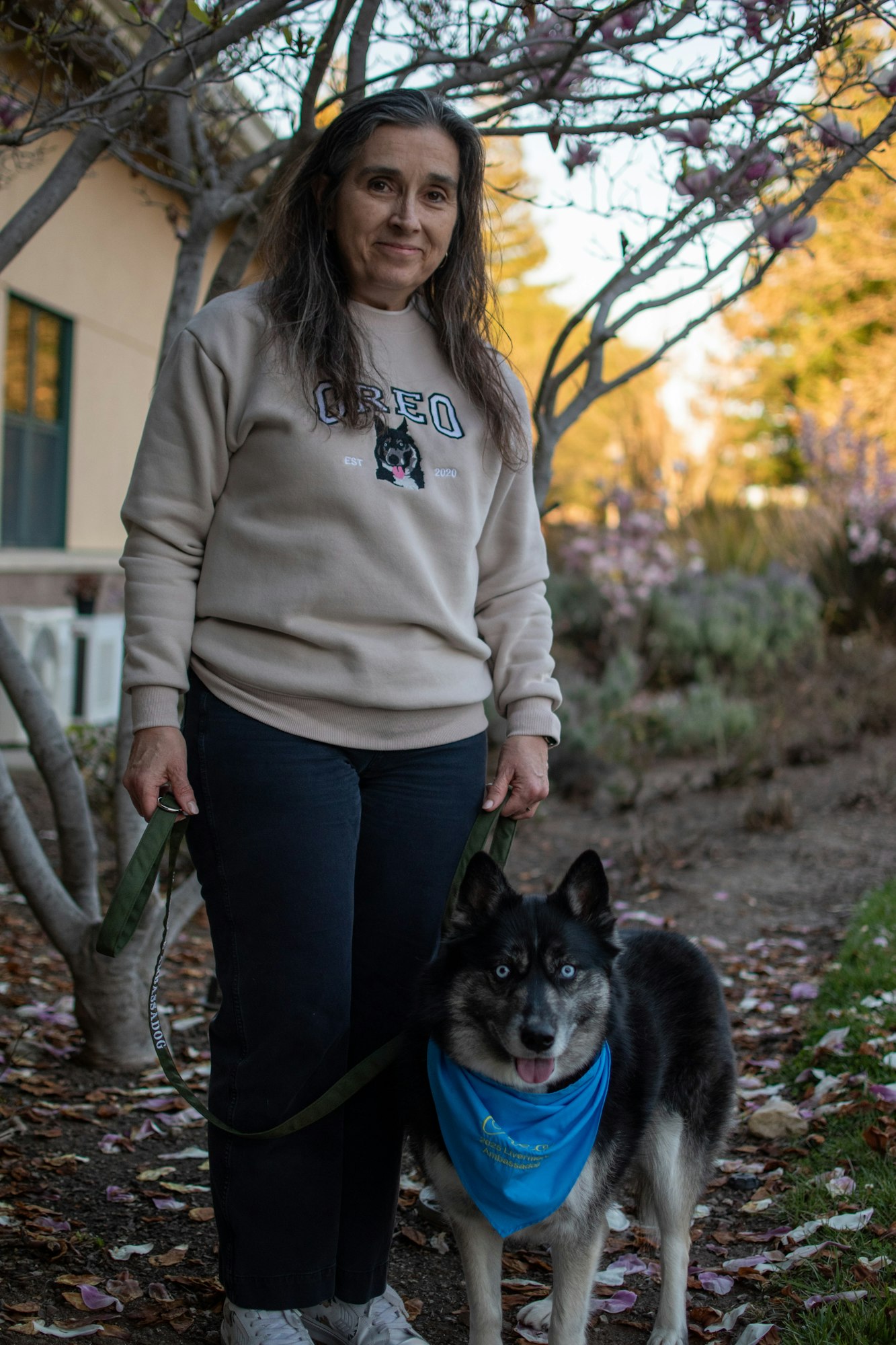 A person with long hair stands outdoors holding a dog on a leash. The dog wears a blue bandana.