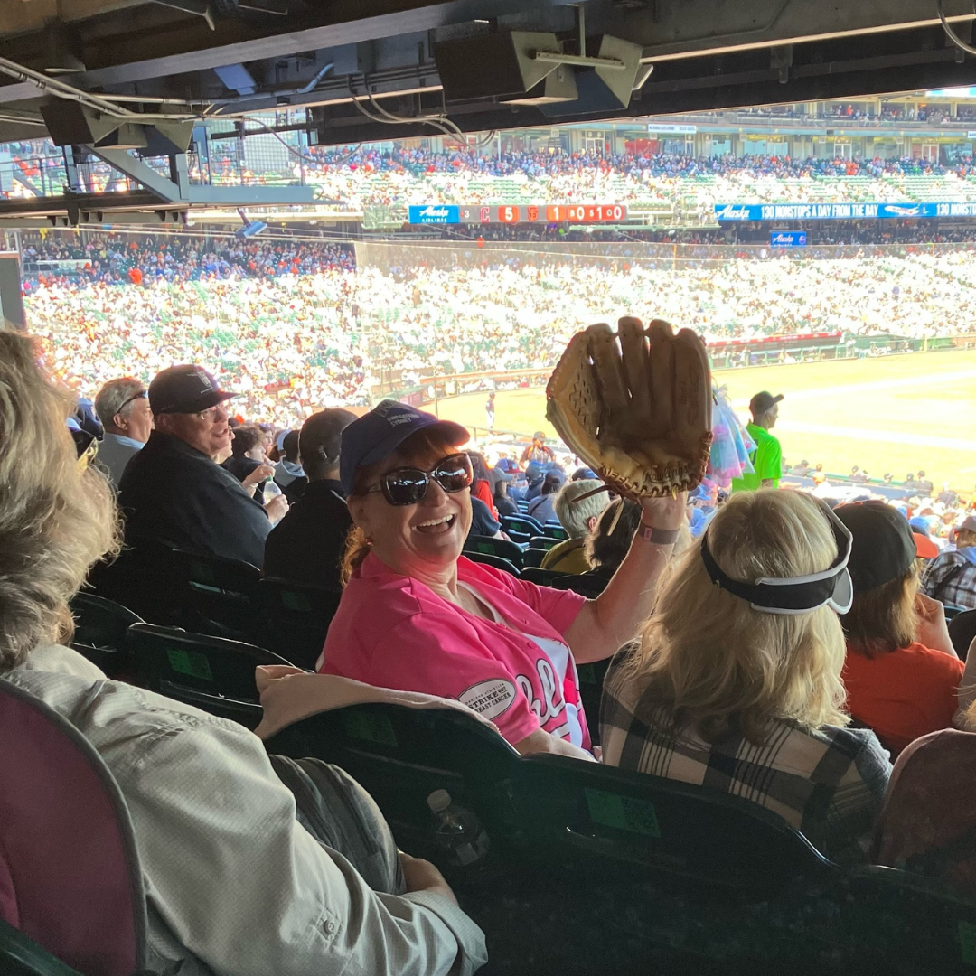 A person in a baseball cap and sunglasses, smiling and holding up a baseball glove at a crowded stadium.