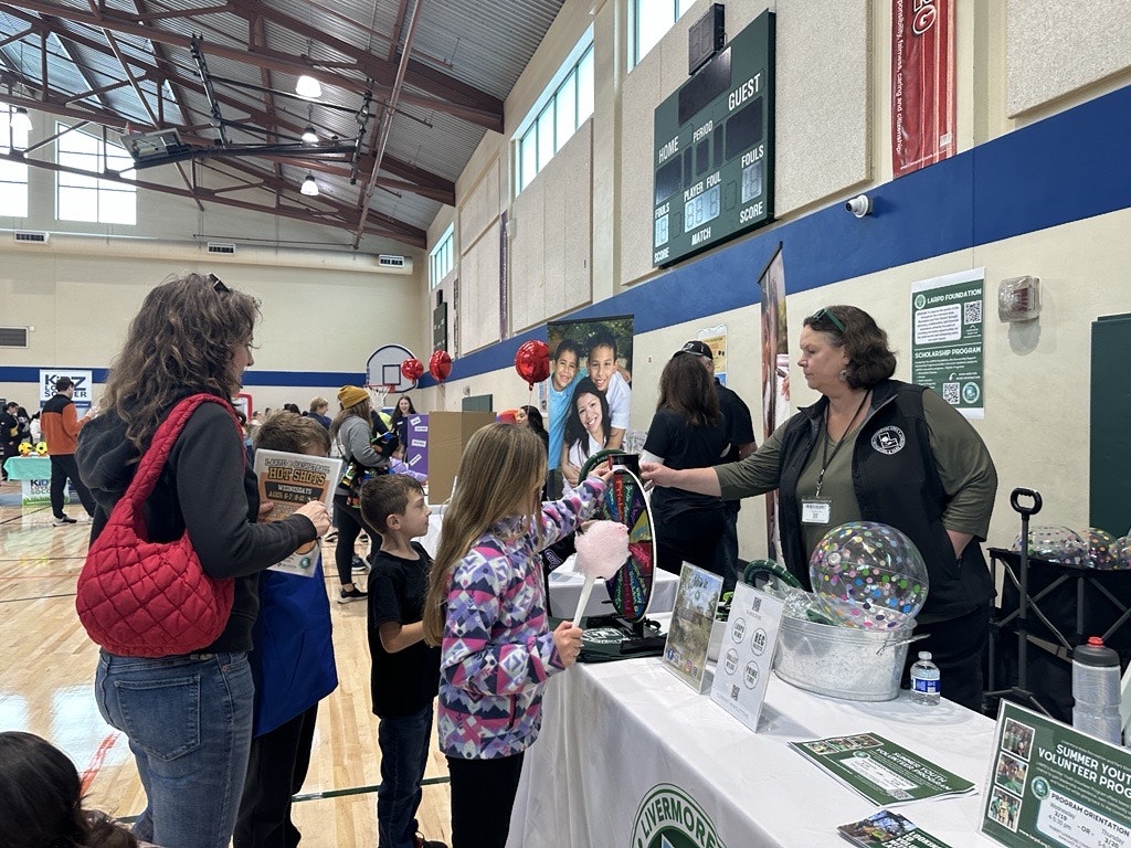 A community event in a gym with families, kids interacting at booths, and a woman serving cotton candy.