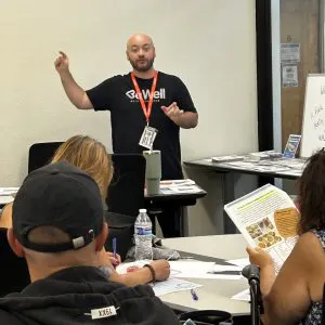 A speaker leads a discussion in a classroom setting, with attendees engaged and taking notes. Materials are visible on the table.