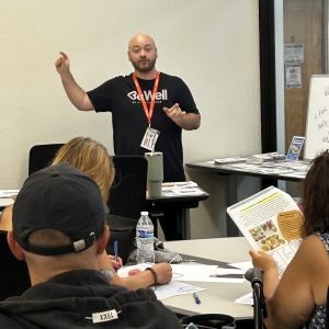 A speaker leads a discussion in a classroom setting, with attendees engaged and taking notes. Materials are visible on the table.
