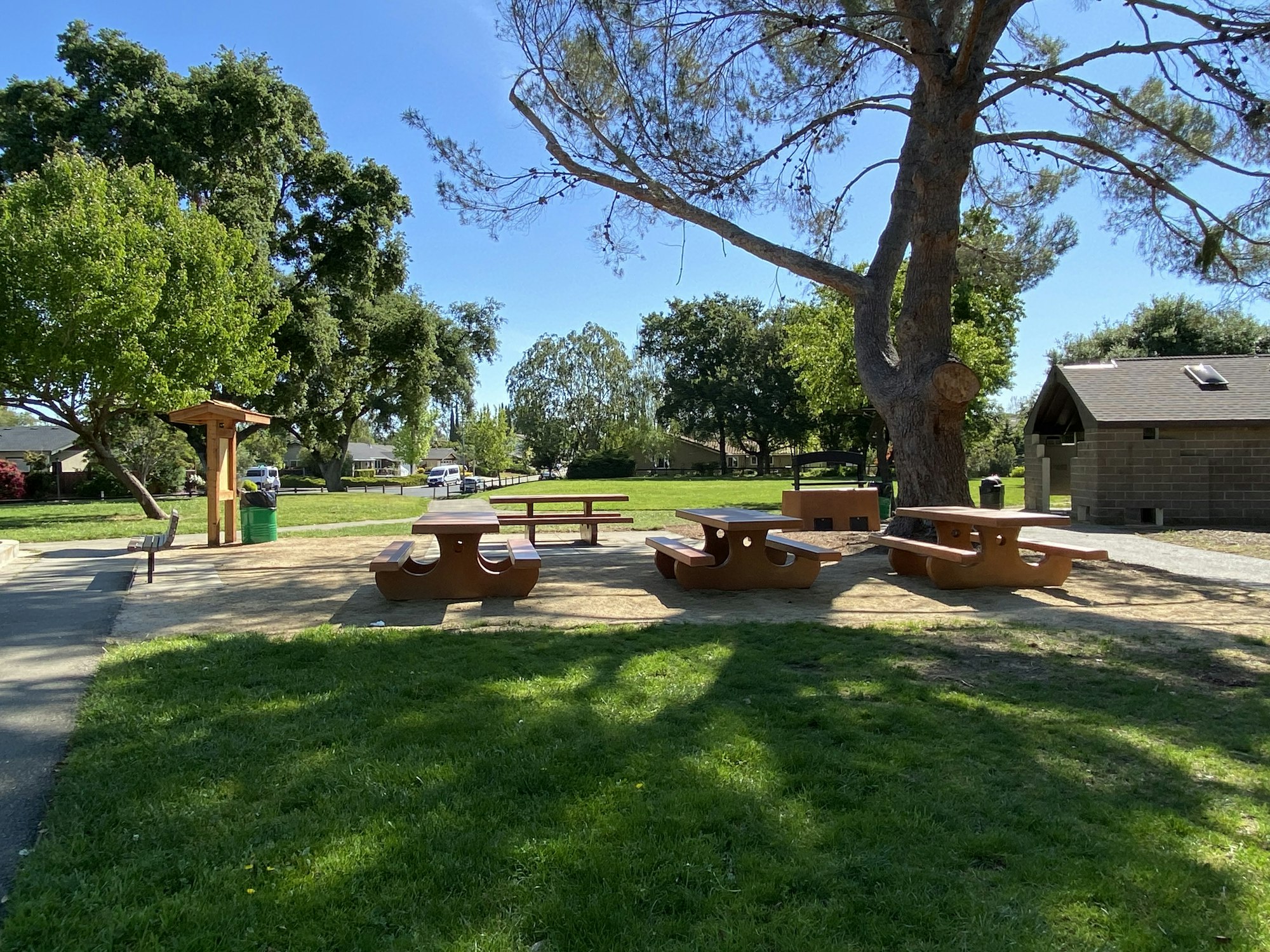 A sunny park with picnic tables, a trash bin, trees, and a restroom building.