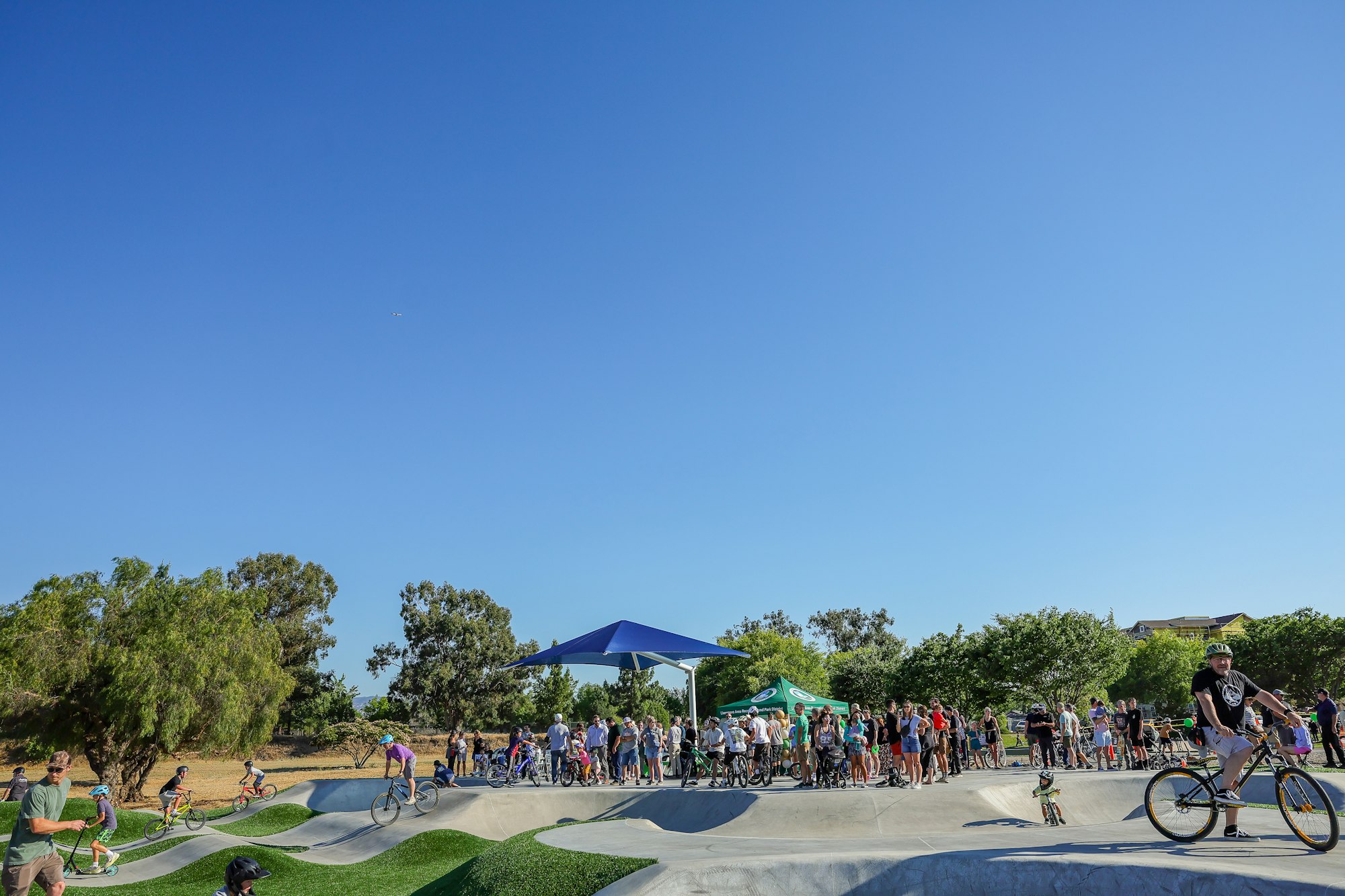 A busy skate park scene with kids riding bikes and a crowd gathered under a canopy, set against a clear blue sky.