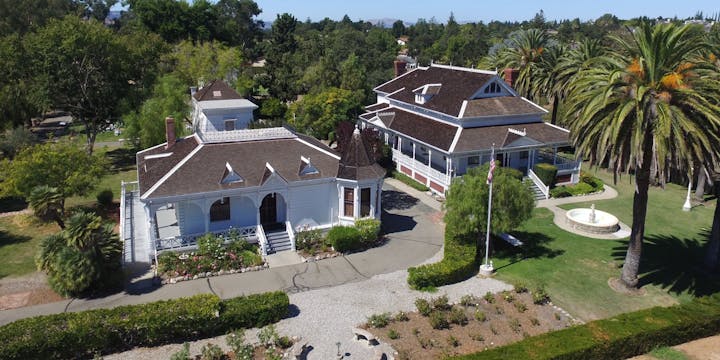 Two large Victorian-style houses surrounded by greenery, palm trees, and a small fountain.