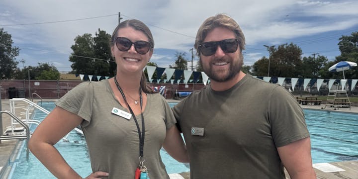 Two smiling individuals in casual attire stand by a swimming pool, with flags and outdoor seating visible in the background.