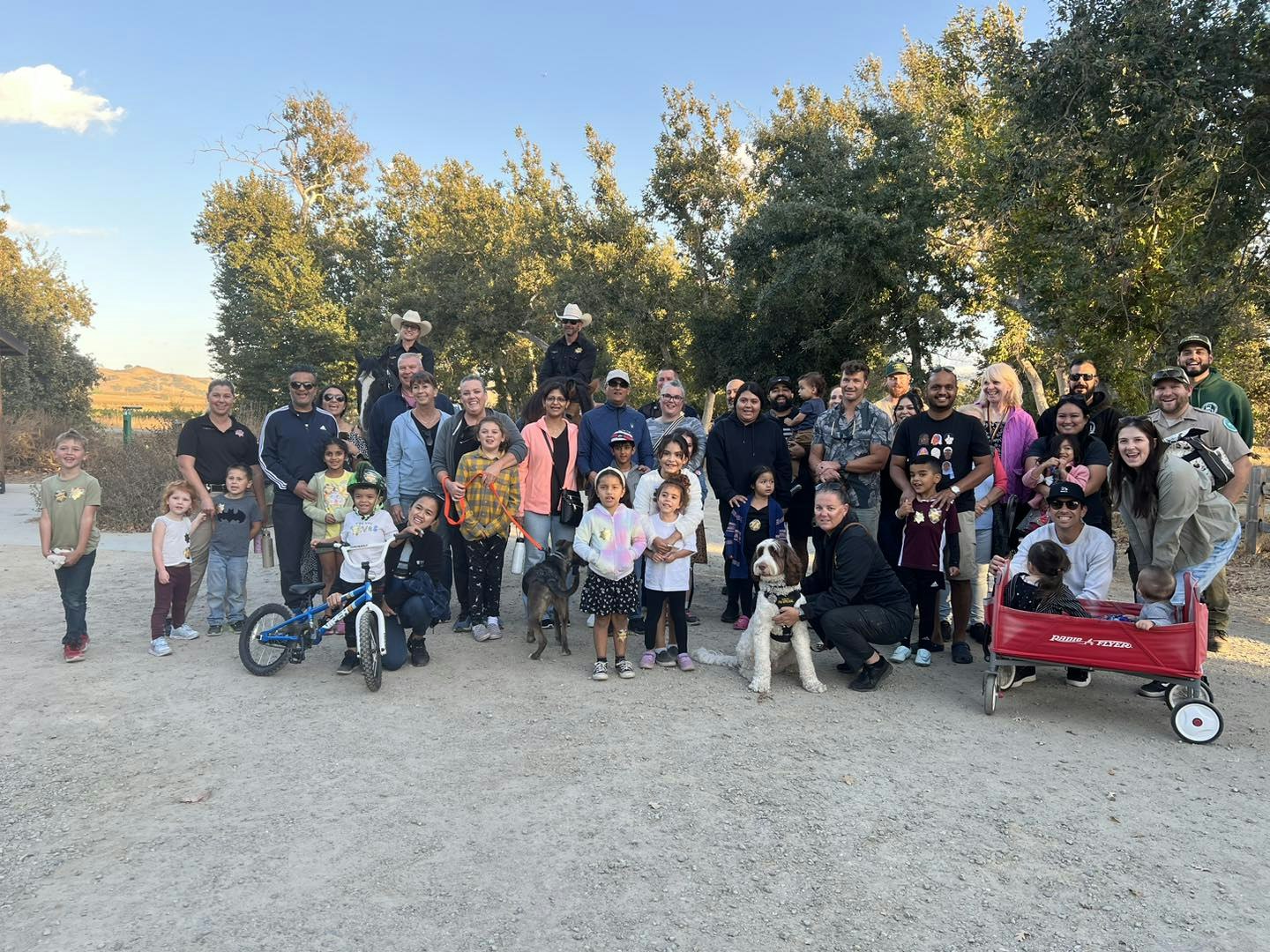 A large group of people and children with dogs, a bike, and a red wagon, gathered outdoors with trees in the background.