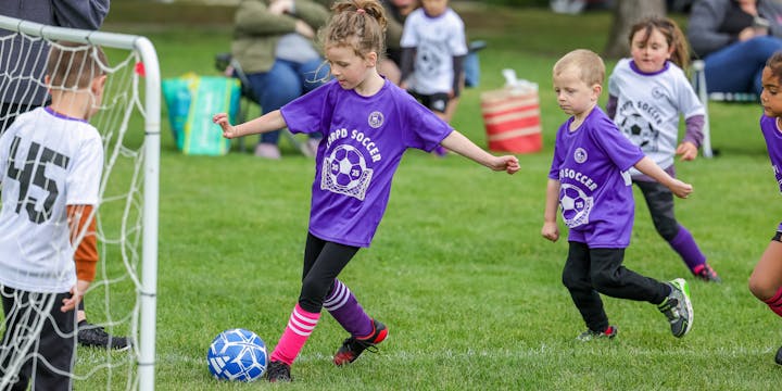 A youth soccer game in progress, featuring kids wearing purple and white jerseys, one girl about to kick the ball.