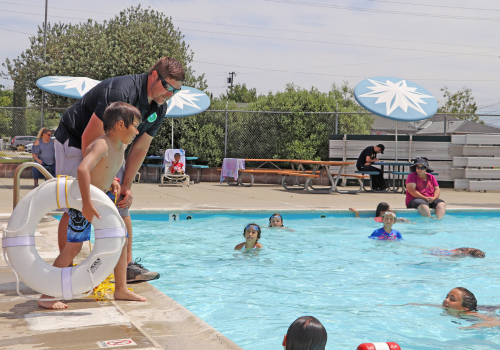 A lifeguard watching a child by a pool, people swimming, loungers, sunny day.
