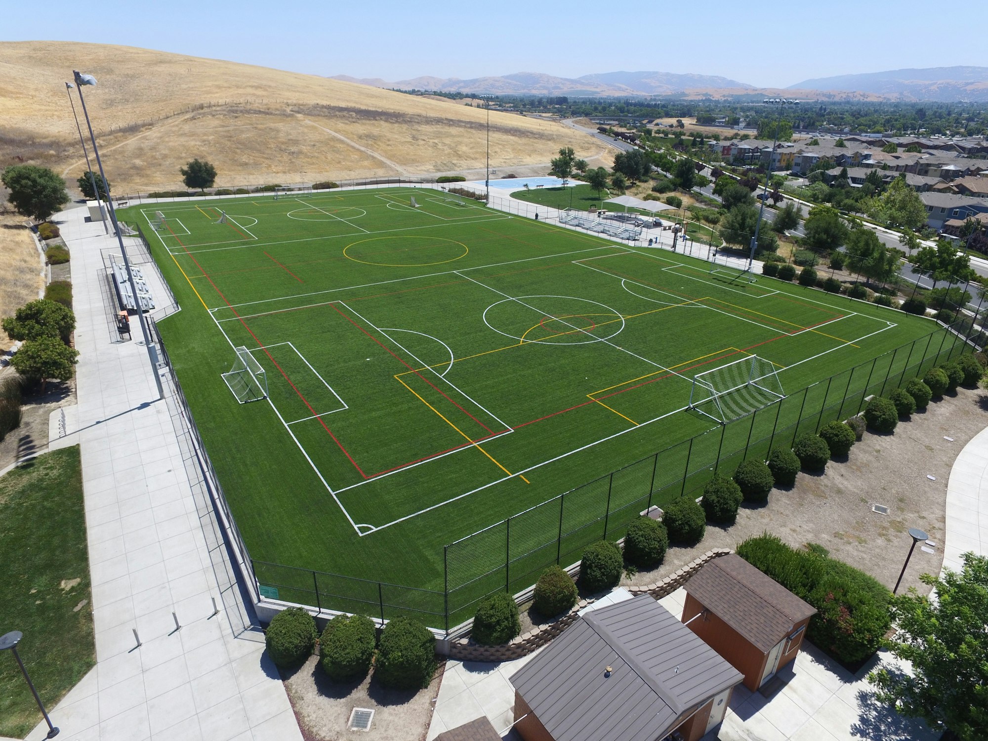 Aerial view of a multi-sport artificial turf field with a soccer pitch, surrounded by fencing, adjacent to a residential area.