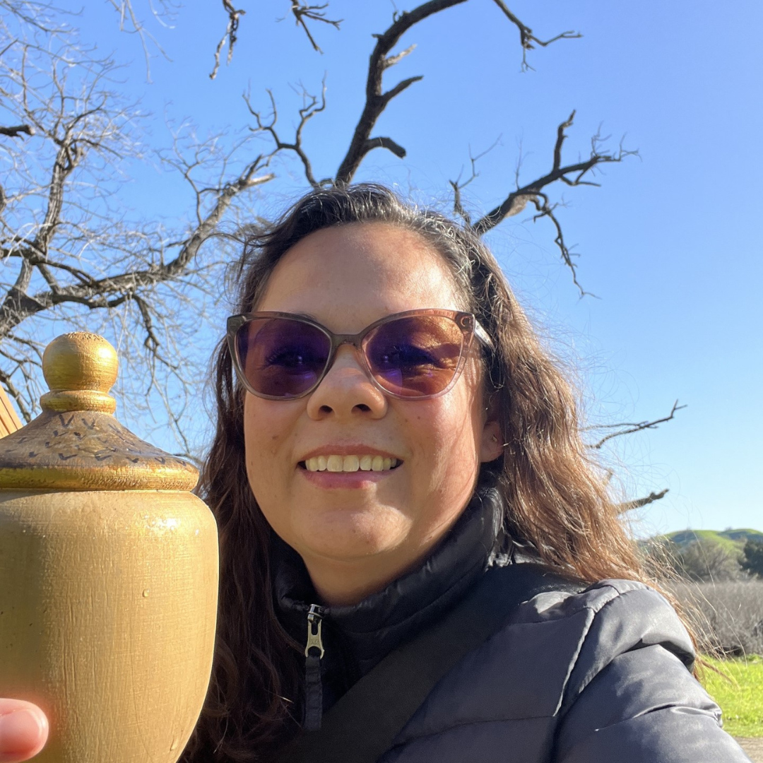 A smiling person with sunglasses holding a jar outdoors, with leafless trees and a blue sky in the background.