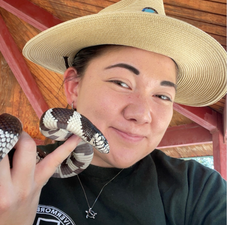 Person wearing a wide-brimmed hat, smiling, holding a black and white snake, under a wooden shelter.