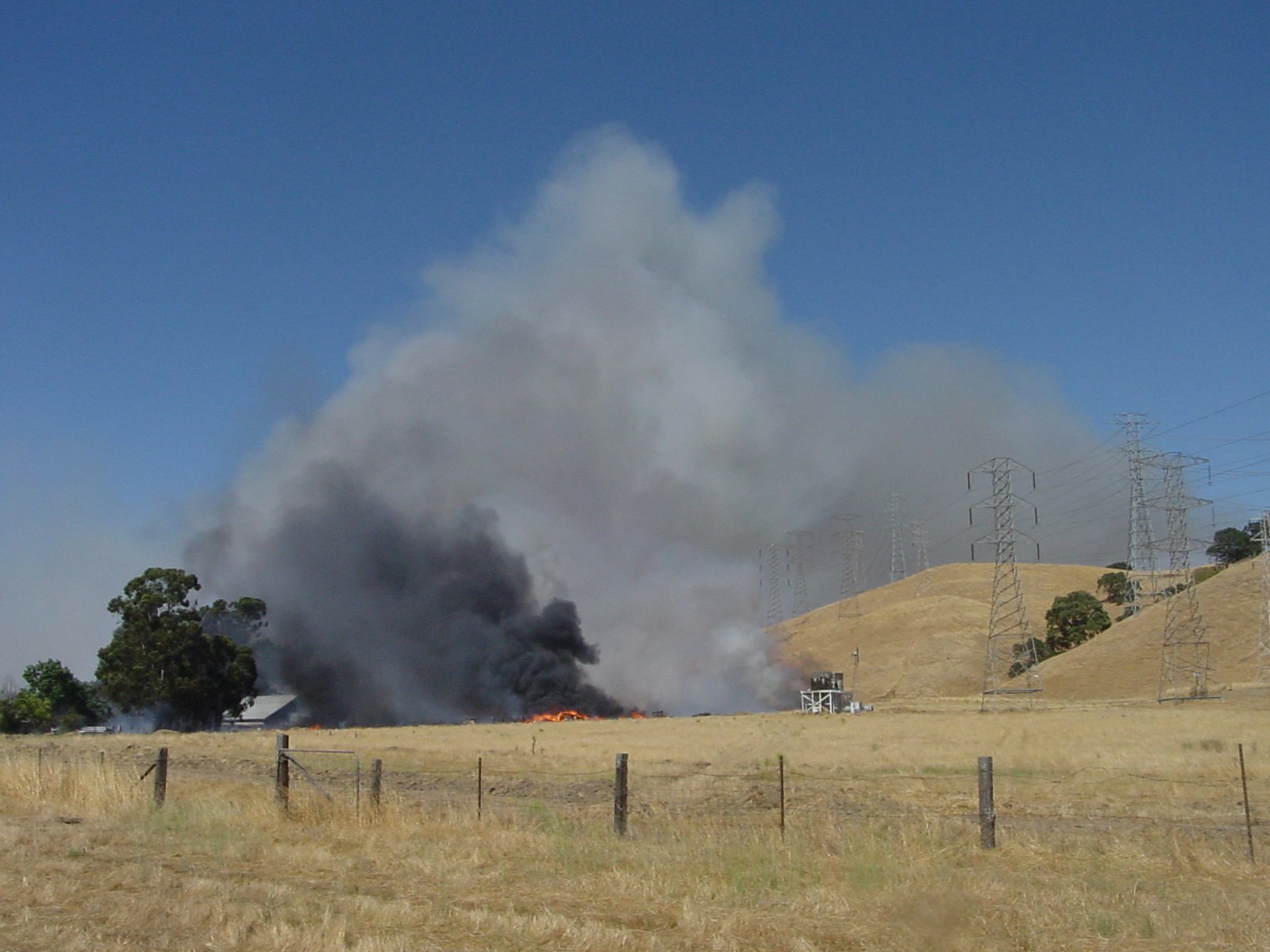 A fire burns the dry, golden grasses at Holdener park near a building; black and white smoke billows back over the hills.