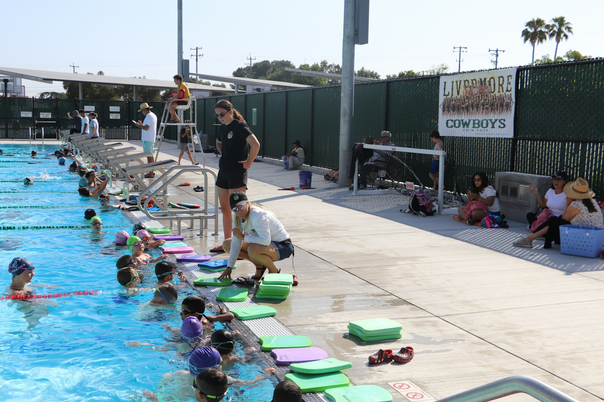 People at a swimming pool, including swimmers with caps and kickboards, coaches, and spectators sitting poolside.