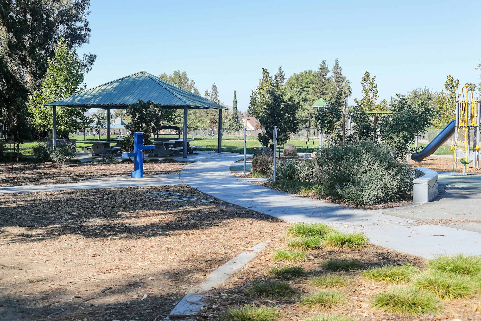 A sunny playground with a slide, swings, picnic tables under a shelter, trees, and a walking path.