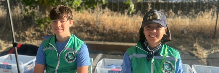 Two individuals wearing green vests and caps are sitting outdoors, likely at a community or recreational event, with storage bins nearby.