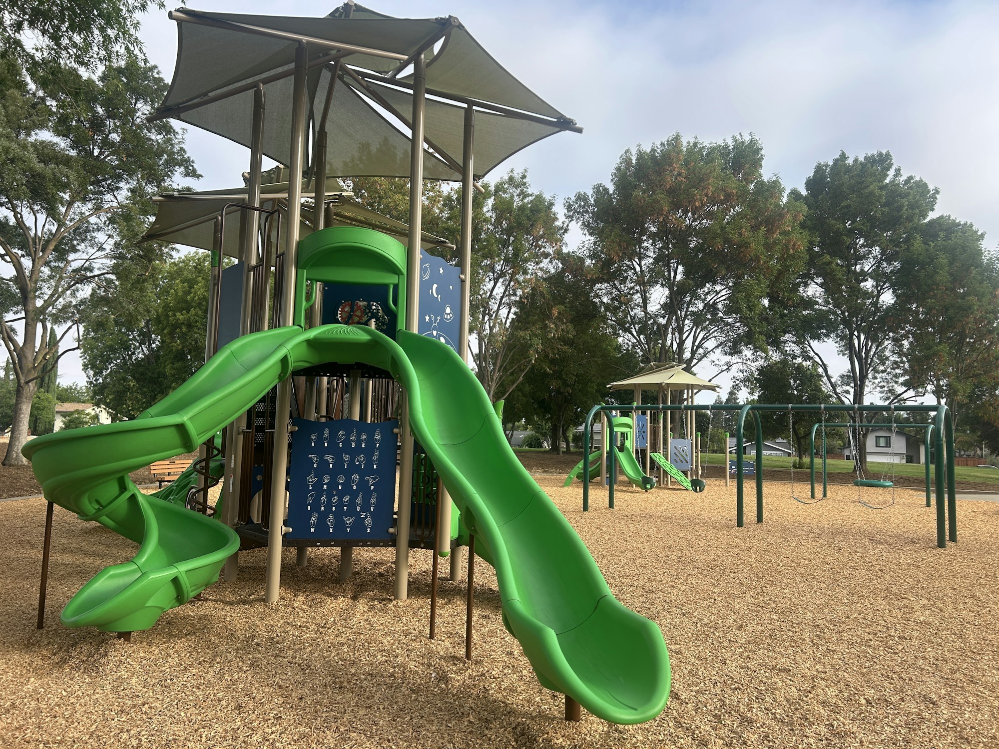 Playground with green slides, swings, climbing structures, and wood chip ground cover.