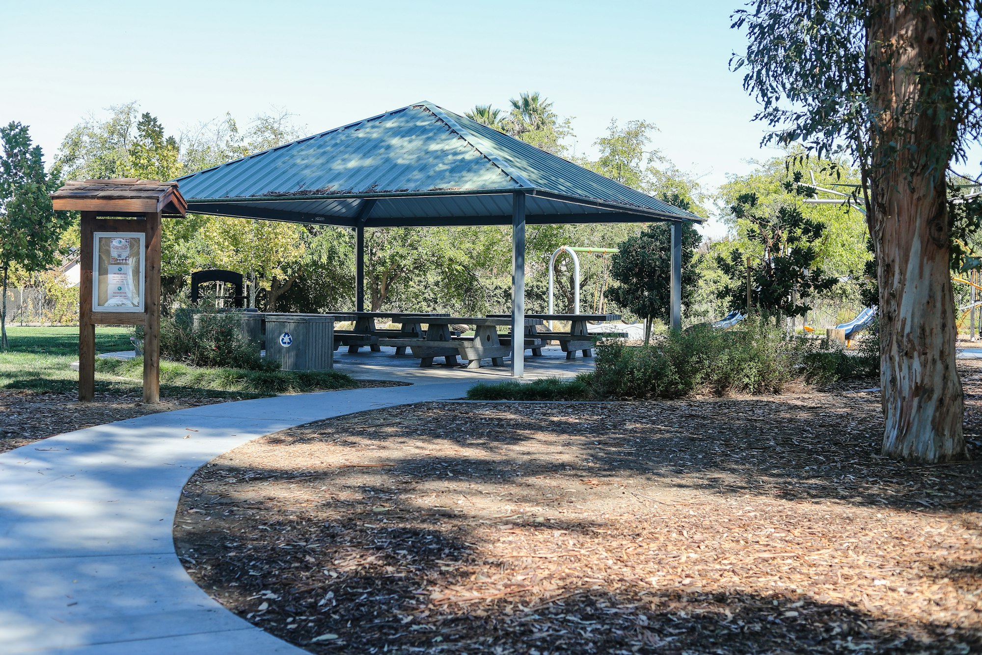 A park pavilion with picnic tables, an information board, a trash can, and trees around.
