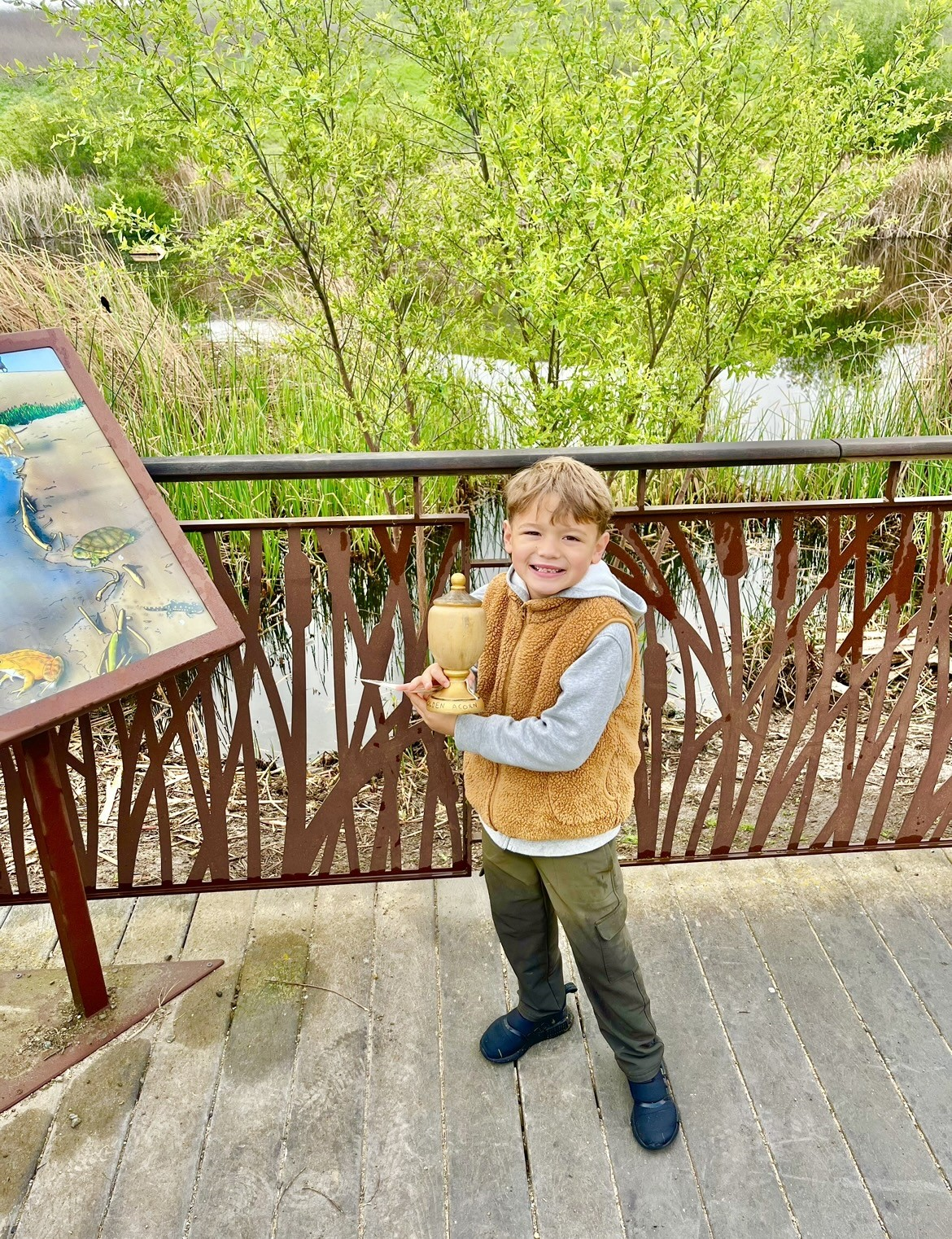 A smiling child holding a cup, standing on a boardwalk with greenery in the background.