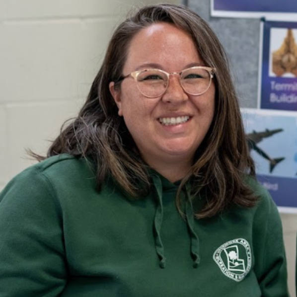 A smiling person wearing glasses and a green hoodie, seated in an indoor setting with educational materials in the background.