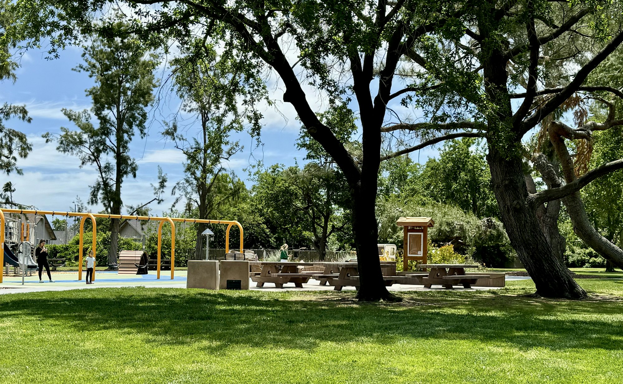Lush park with trees, grass, playground, benches, and people enjoying the outdoors.