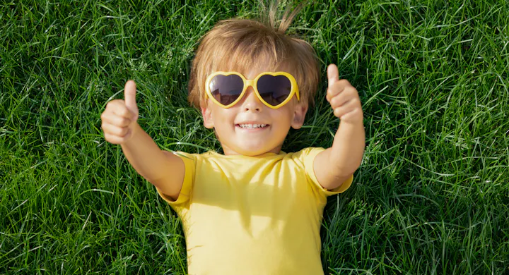 A smiling child wearing yellow heart-shaped sunglasses and a yellow shirt, lying on green grass and giving a thumbs up.